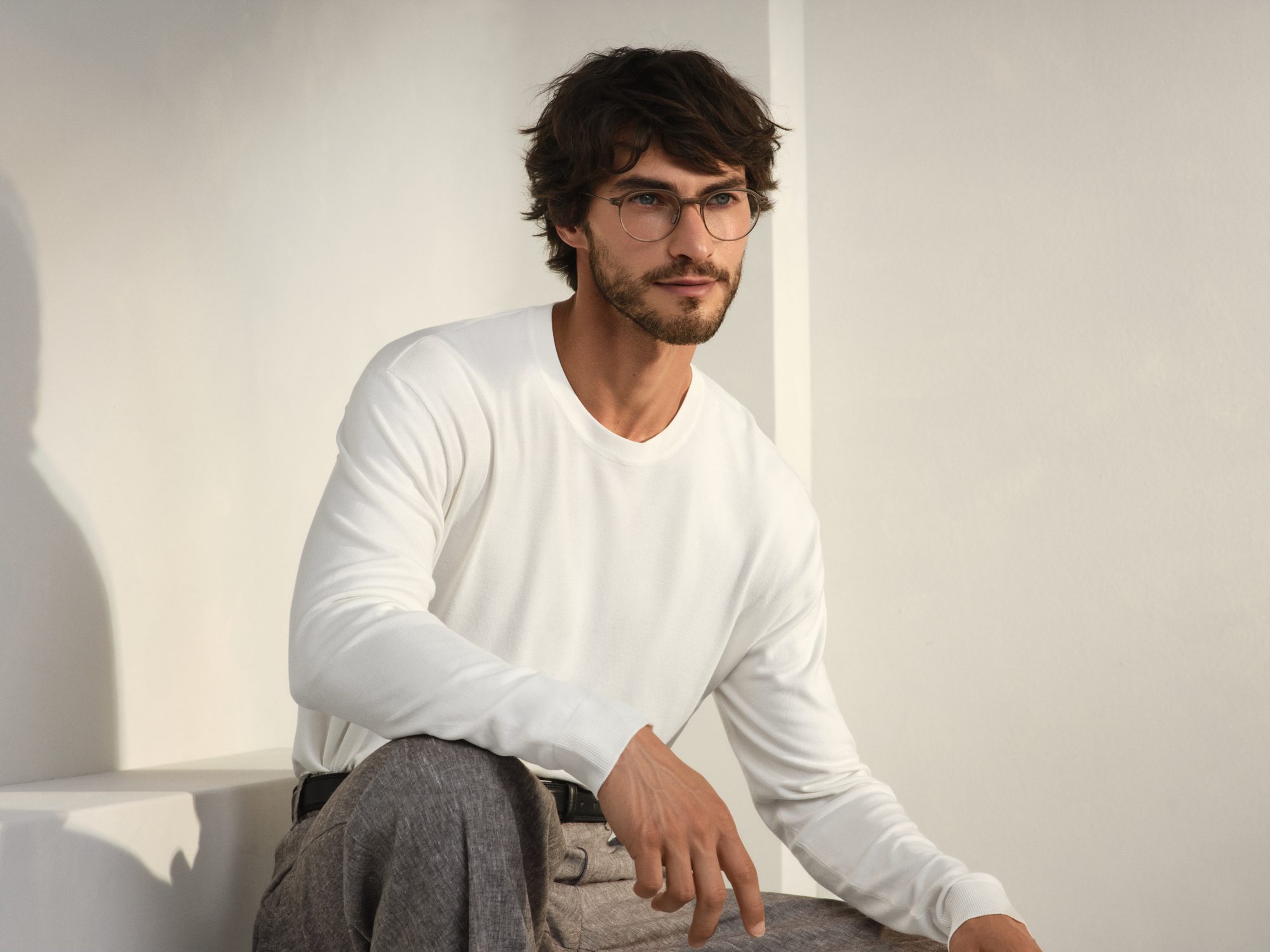Bearded man in a white sweater wearing Rodenstock eyeglasses, seated by a wall