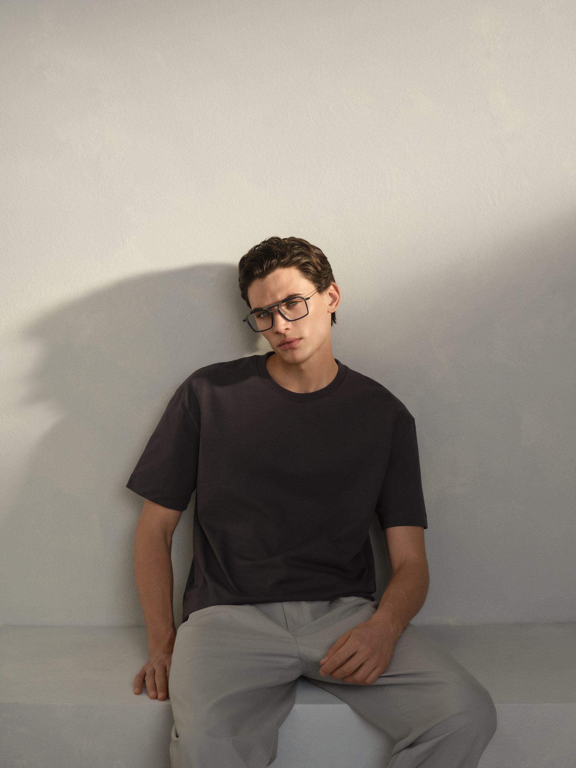 Man in a black T-shirt wearing dark Rodenstock frames, leaning against a light wall