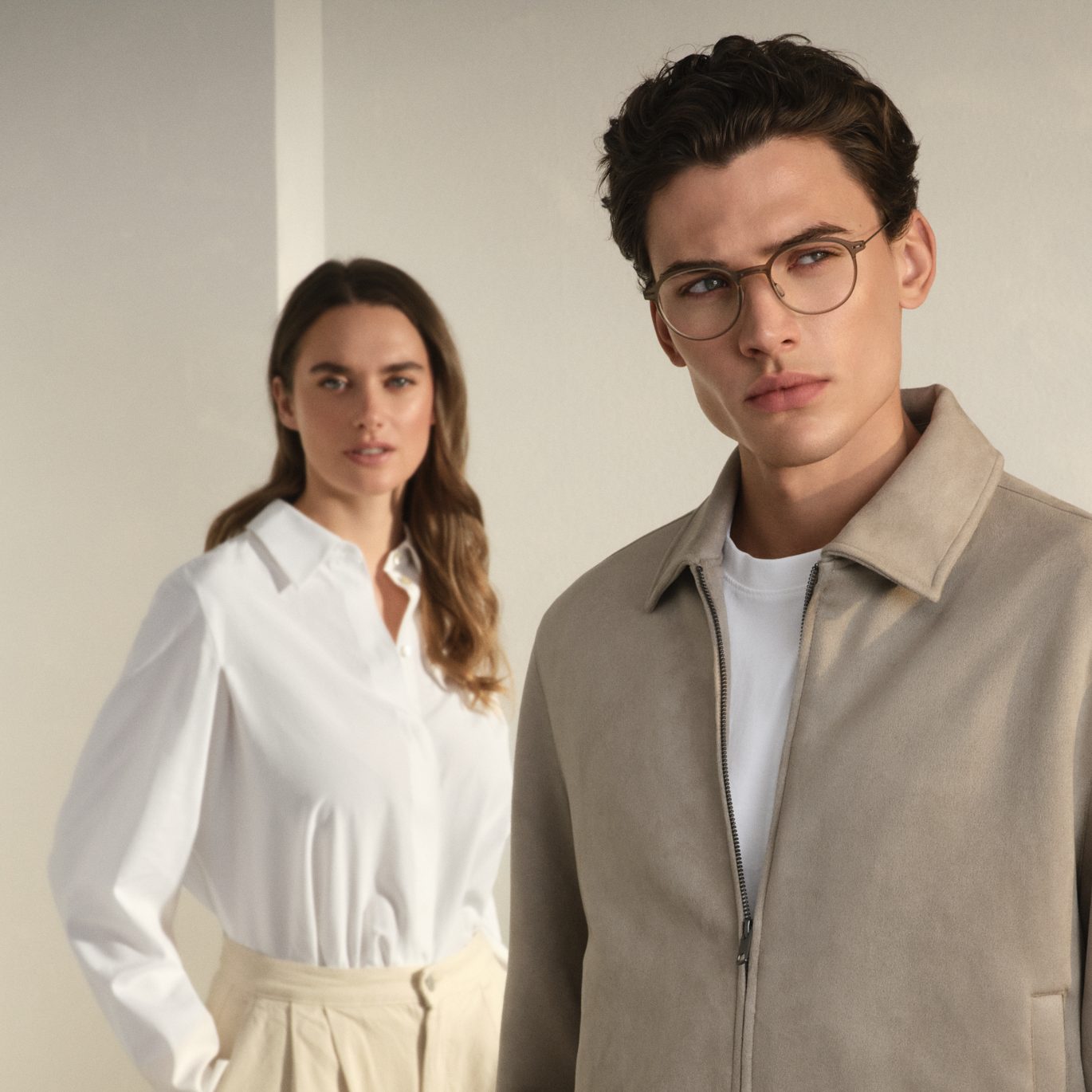 Man in a beige jacket wearing round Rodenstock eyeglasses, a woman in a white blouse in the background